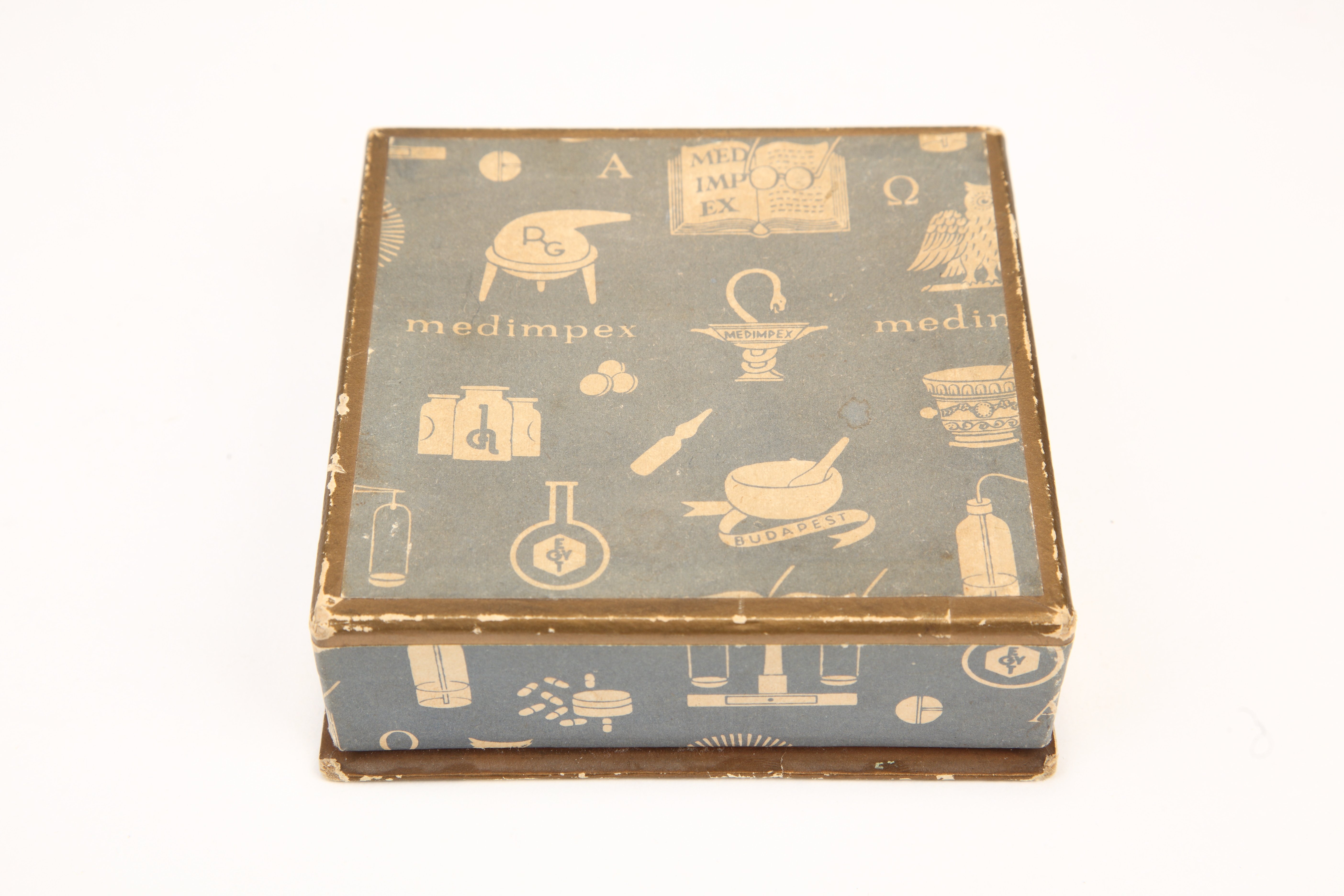 Négyzet alakú arany szélű fedeles világoskék alapszínű doboz. Felül és oldalt gyógyszergyári szimbólumok és rajzok: üveg, laboreszköz, kön (Magyar Kereskedelmi és Vendéglátóipari Múzeum CC BY-NC-SA)